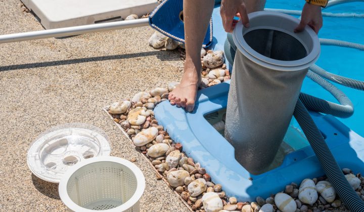 A person removing a used filter cartridge from a pool filtration unit, with the replacement cartridge and lid nearby on a pebble-lined deck.